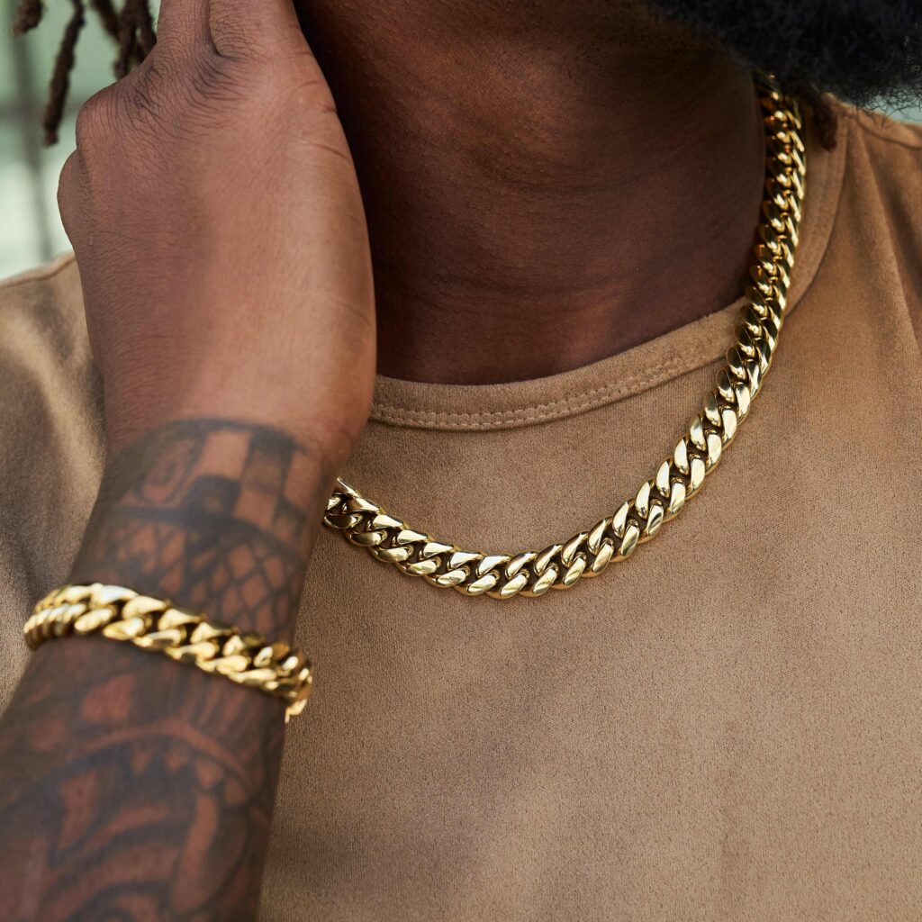 Close-up of a man wearing a gold Cuban link chain necklace and matching bracelet with a tattooed arm visible.